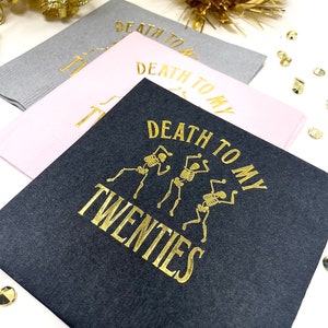 May include: Three square cocktail napkins in gray, pink, and black. The black napkin features gold text and skeleton illustrations that read "Death to my Twenties." The pink and gray napkins have the text "Death to my" in gold. Gold confetti and tinsel are in the background.