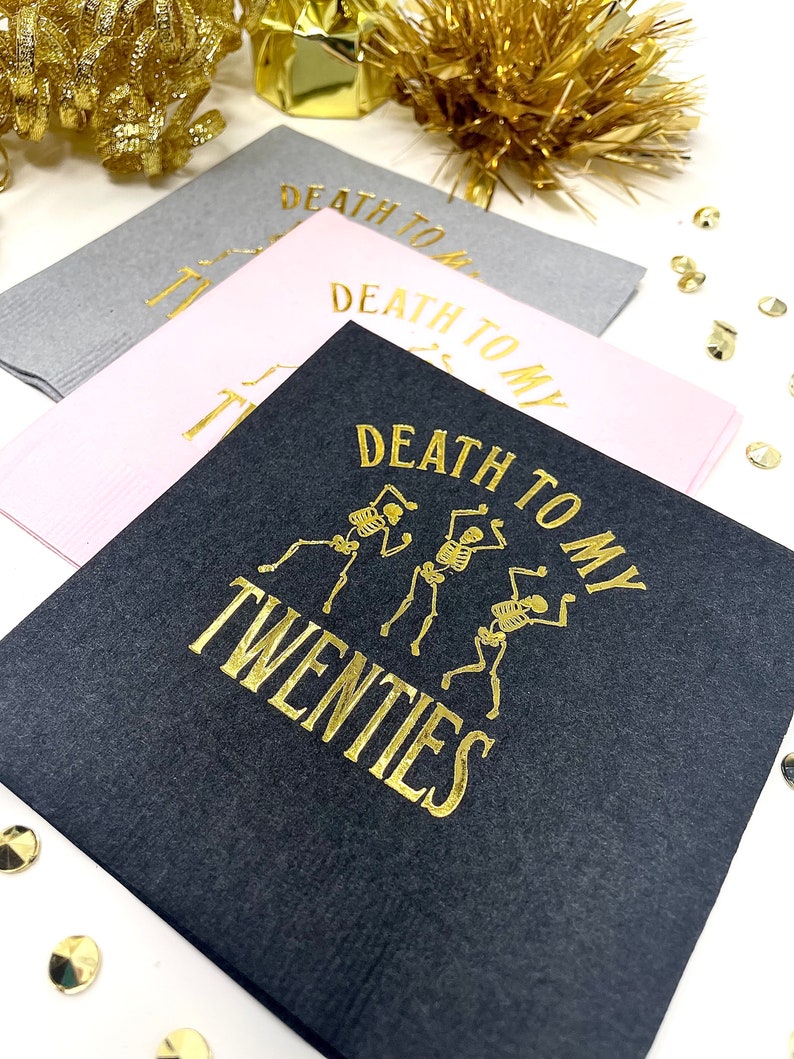 May include: Three square cocktail napkins in gray, pink, and black. The black napkin features gold text and skeleton illustrations that read "Death to my Twenties." The pink and gray napkins have the text "Death to my" in gold. Gold confetti and tinsel are in the background.