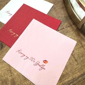 Könnte beinhalten: Drei quadratische Papierservietten in Weiß, Rot und Rosa. Die rosa Serviette trägt den Text "Kissing my 20's Goodbye" mit einer Lippen-Grafik. Die roten und weißen Servietten tragen ebenfalls den Text "Kissing my 20's Goodbye".