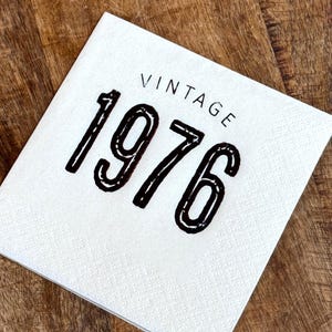 May include: A white square napkin with the word "VINTAGE" printed above the number "1976" in black. The napkin is on a wooden surface.