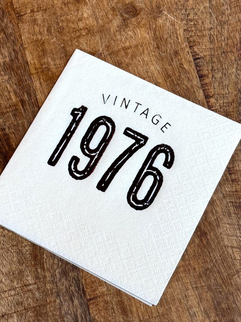 May include: A white square napkin with the word "VINTAGE" printed above the number "1976" in black. The napkin is on a wooden surface.