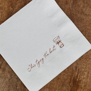 A single white folded square napkin with "She's Tying the Knot" and a small bow illustration printed in rose gold foil.