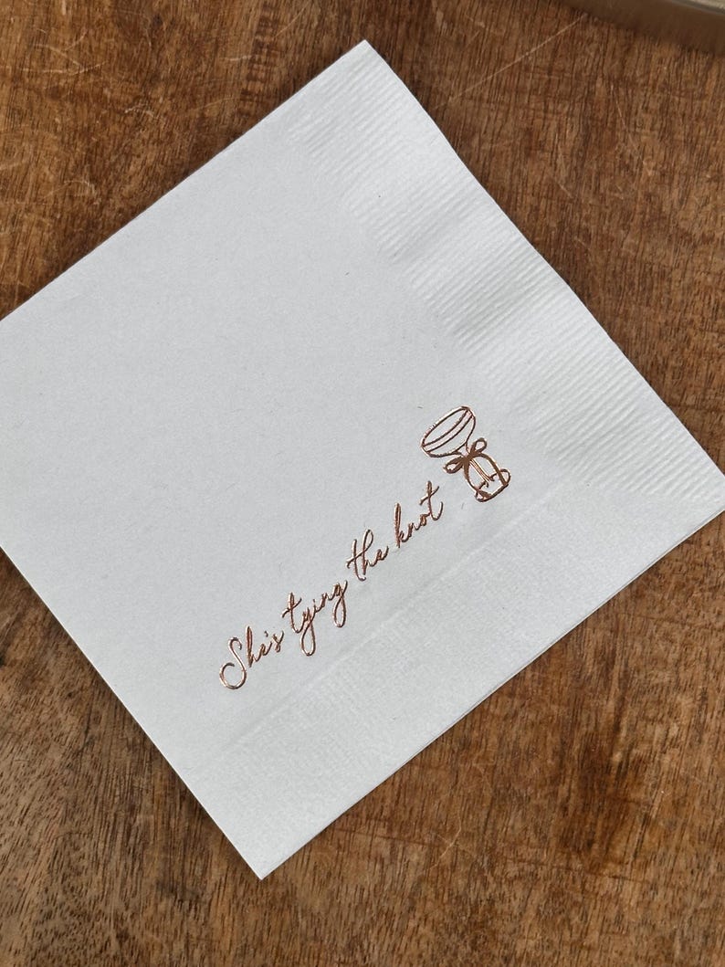 A single white folded square napkin with "She's Tying the Knot" and a small bow illustration printed in rose gold foil.