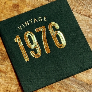 May include: A dark green square napkin with the word "VINTAGE" above the gold metallic numbers "1976". The napkin is on a wooden surface. The napkin is likely for a birthday or anniversary celebration.