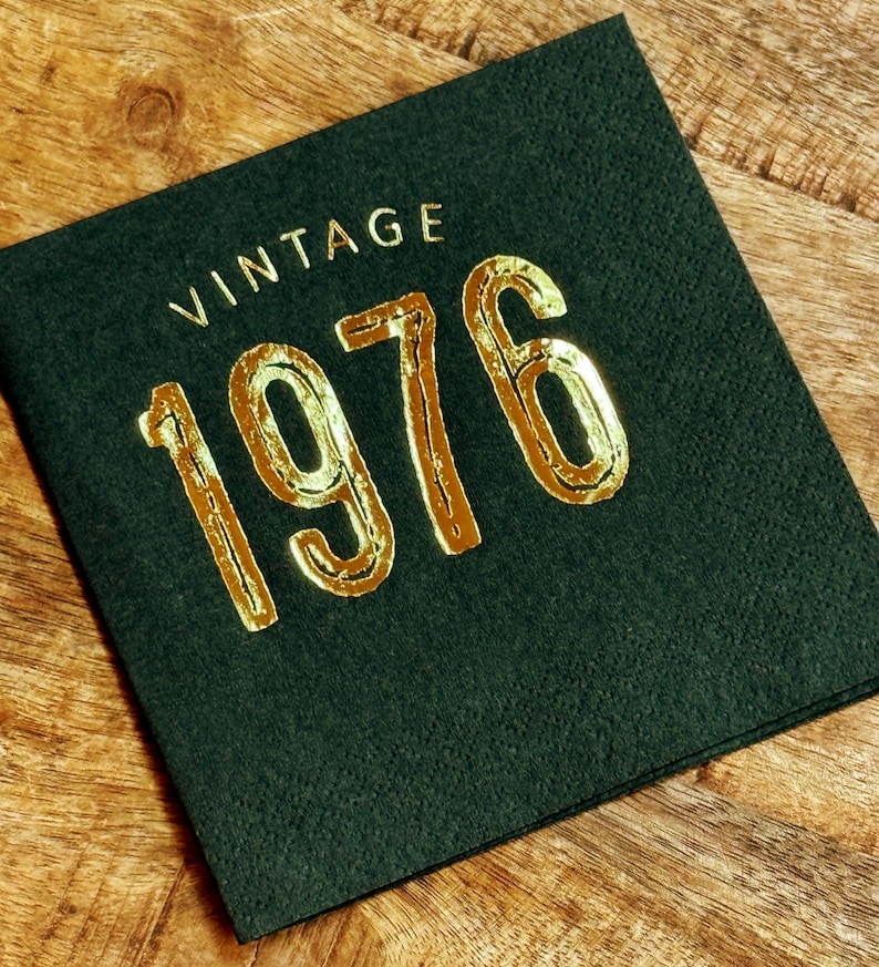May include: A dark green square napkin with the word "VINTAGE" above the gold metallic numbers "1976". The napkin is on a wooden surface. The napkin is likely for a birthday or anniversary celebration.