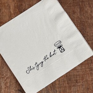 A single white folded square napkin with "She's Tying the Knot" and a small bow illustration printed in black ink.