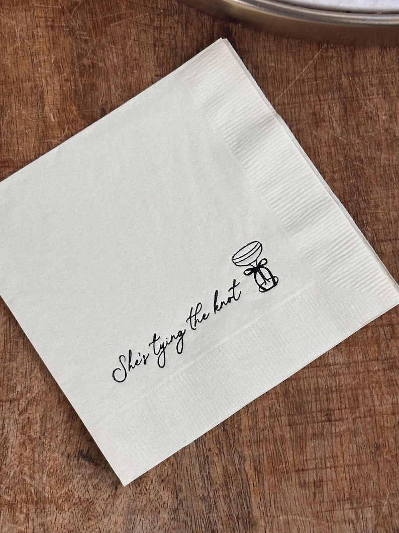 A single white folded square napkin with "She's Tying the Knot" and a small bow illustration printed in black ink.