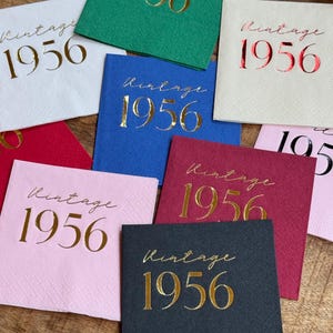 May include: A collection of square paper napkins in various colors, including white, green, blue, red, pink, and black. Each napkin features the gold foil text "Vintage 1956" in a script font, suitable for a birthday celebration.