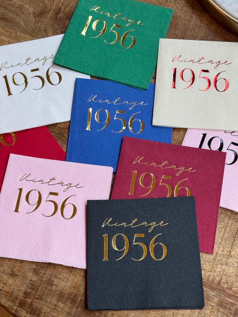 May include: A collection of square paper napkins in various colors, including white, green, blue, red, pink, and black. Each napkin features the gold foil text "Vintage 1956" in a script font, suitable for a birthday celebration.