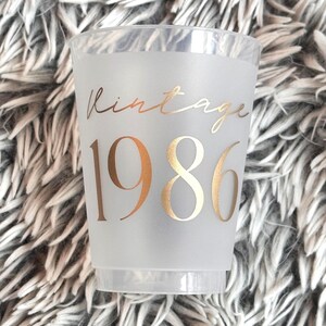 A single frosted plastic cup with "vintage" in gold script and "1986" in gold block letters is shown on a fuzzy gray background.