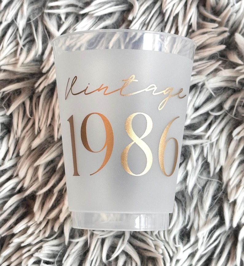 A single frosted plastic cup with "vintage" in gold script and "1986" in gold block letters is shown on a fuzzy gray background.