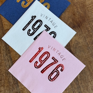 May include: Three square napkins in blue, white, and pink, each with the word "VINTAGE" above a year. The blue napkin has "1970" in gold, the white has "1970" in black, and the pink has "1976" in red.