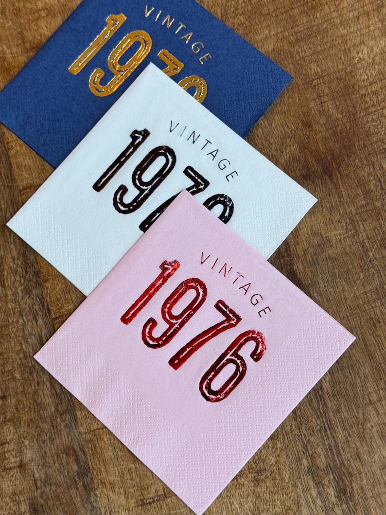May include: Three square napkins in blue, white, and pink, each with the word "VINTAGE" above a year. The blue napkin has "1970" in gold, the white has "1970" in black, and the pink has "1976" in red.