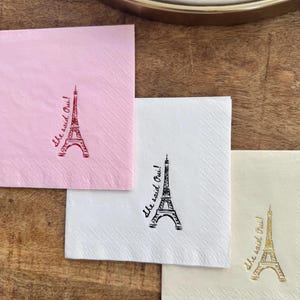 May include: Four paper napkins in pink, white, and cream, each with an Eiffel Tower design and the phrase "She said Oui!". The napkins are arranged on a wooden surface.