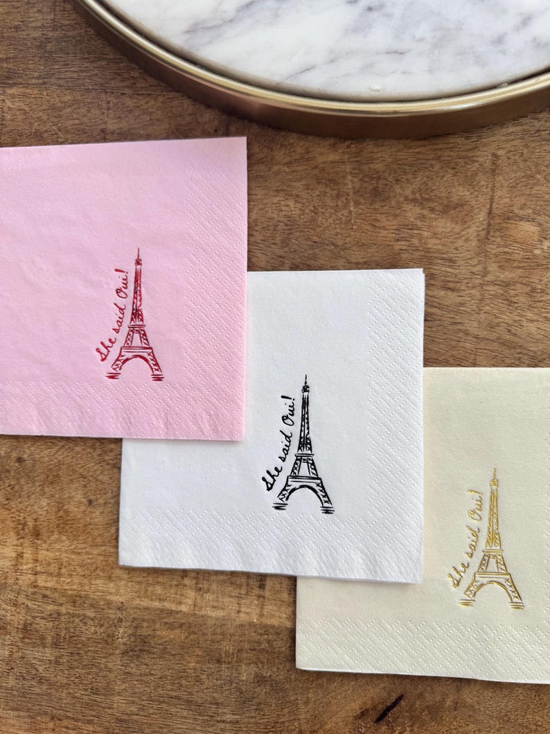 May include: Four paper napkins in pink, white, and cream, each with an Eiffel Tower design and the phrase "She said Oui!". The napkins are arranged on a wooden surface.