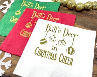 May include: Three square paper napkins in white, red, and green, each featuring gold foil text that reads Ball's Deep in CHRISTMAS CHEER, accompanied by illustrations of four hanging Christmas ornaments and a small snowman face.