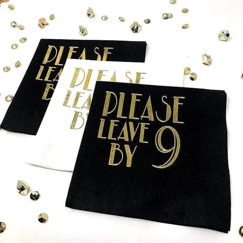 Top-down view of black and white paper napkins with gold foil stamped text saying Please Leave By 9, arranged on a white background with scattered gold foil flakes.