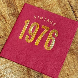 May include: A square, burgundy-colored napkin with the word "VINTAGE" and the number "1976" in gold lettering. The napkin is on a wooden surface.