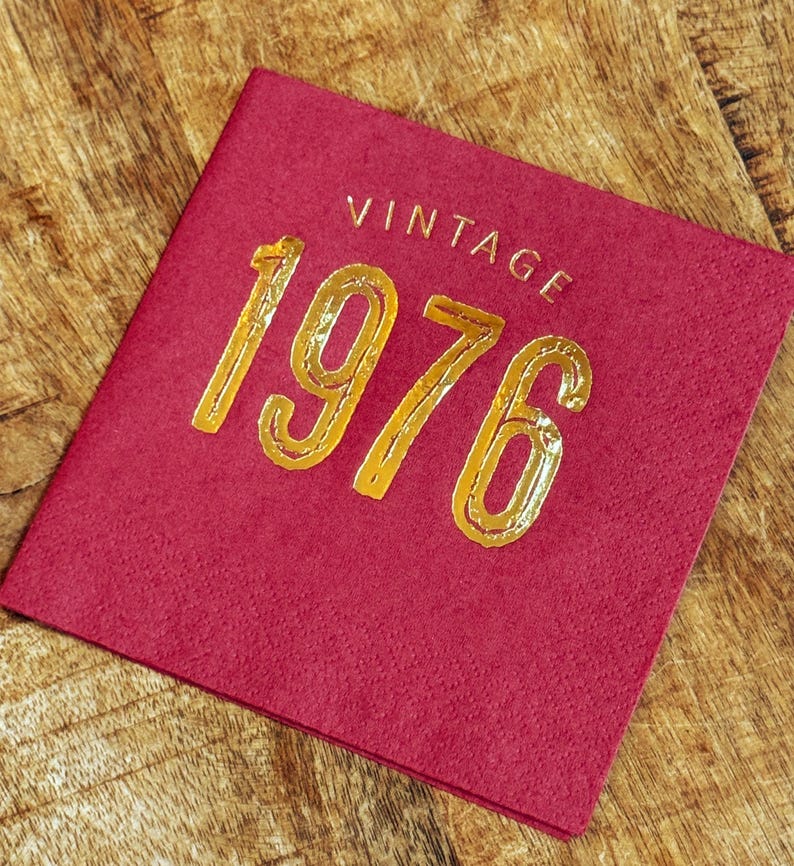 May include: A square, burgundy-colored napkin with the word "VINTAGE" and the number "1976" in gold lettering. The napkin is on a wooden surface.