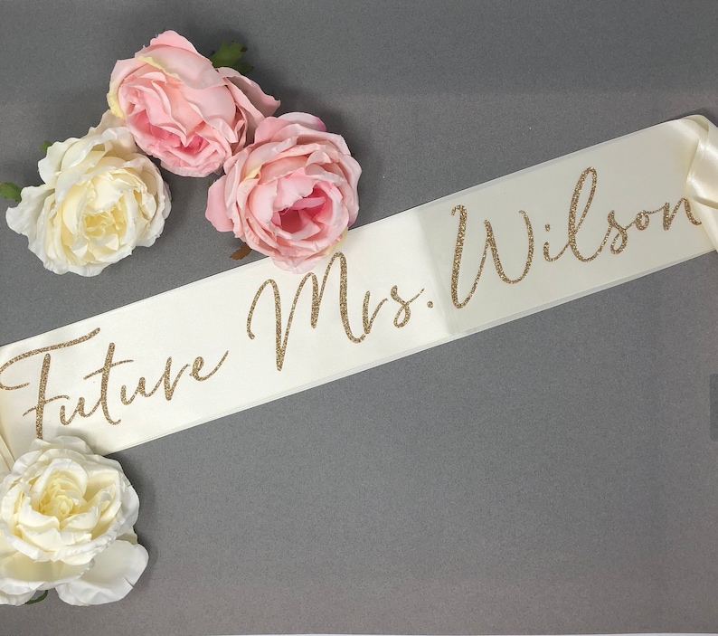 A cream satin sash with gold glitter lettering that reads 'Future Mrs. Wilson'.   The writing can be customized.  Custom Future Mrs. Sash cream with Gold glitter