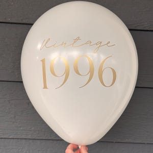 May include: A white balloon with gold text reading "Vintage 1996". The balloon is held by a hand. The background is a grey wooden surface. The balloon is round and inflated.