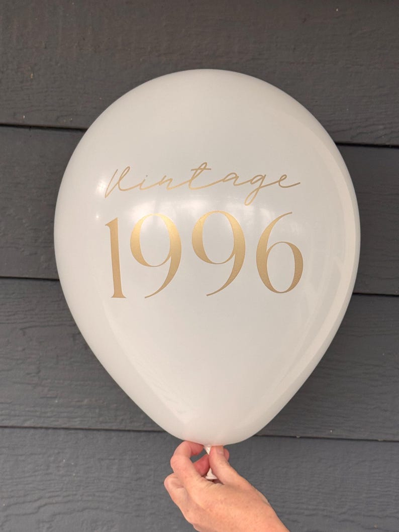 May include: A white balloon with gold text reading "Vintage 1996". The balloon is held by a hand. The background is a grey wooden surface. The balloon is round and inflated.