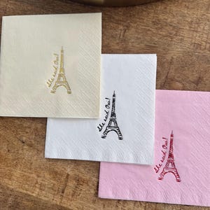 May include: Three square paper napkins in cream, white, and pink, each with a printed Eiffel Tower design and the phrase "She said Oui!". The napkins are on a wooden surface.