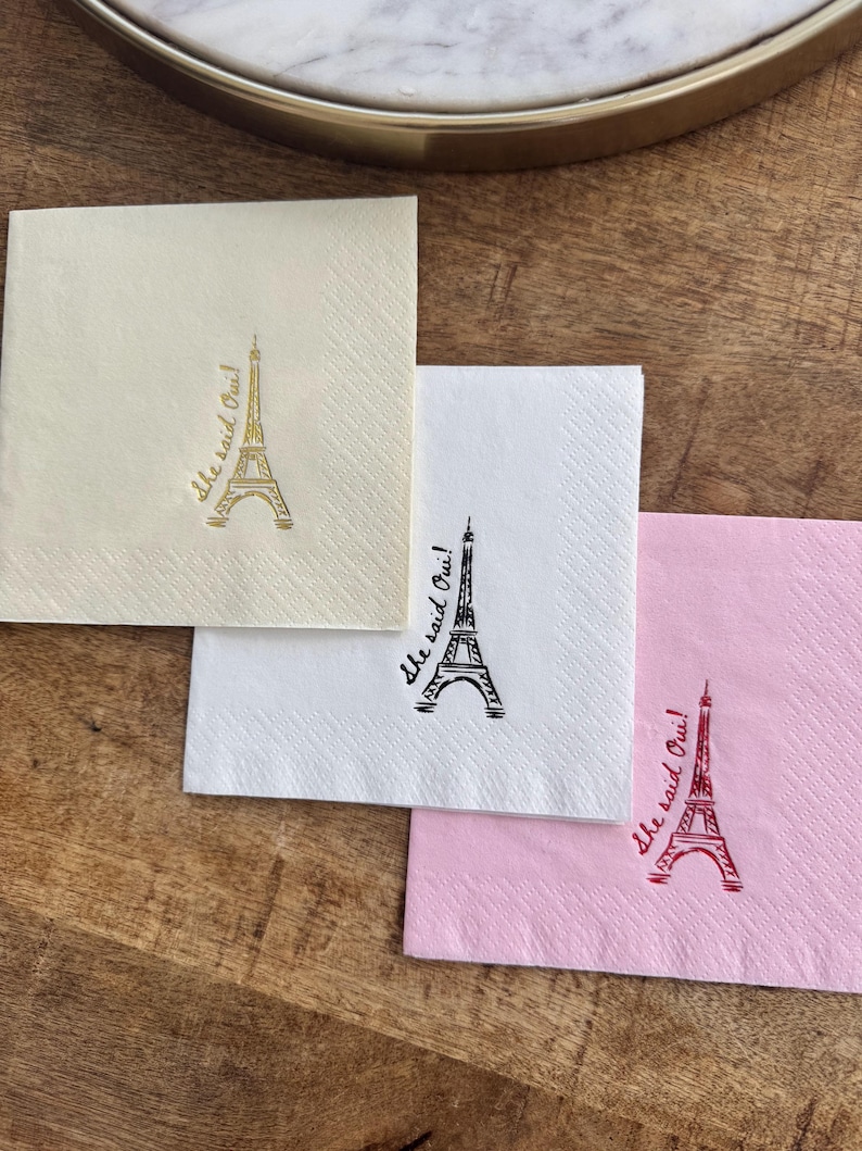 May include: Three square paper napkins in cream, white, and pink, each with a printed Eiffel Tower design and the phrase "She said Oui!". The napkins are on a wooden surface.