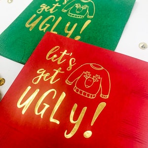 May include: Two square paper napkins, one green and one red, each featuring gold foil text that reads "let's get UGLY!" alongside a simple line drawing of a sweater with a reindeer face on the front, arranged on a white surface.