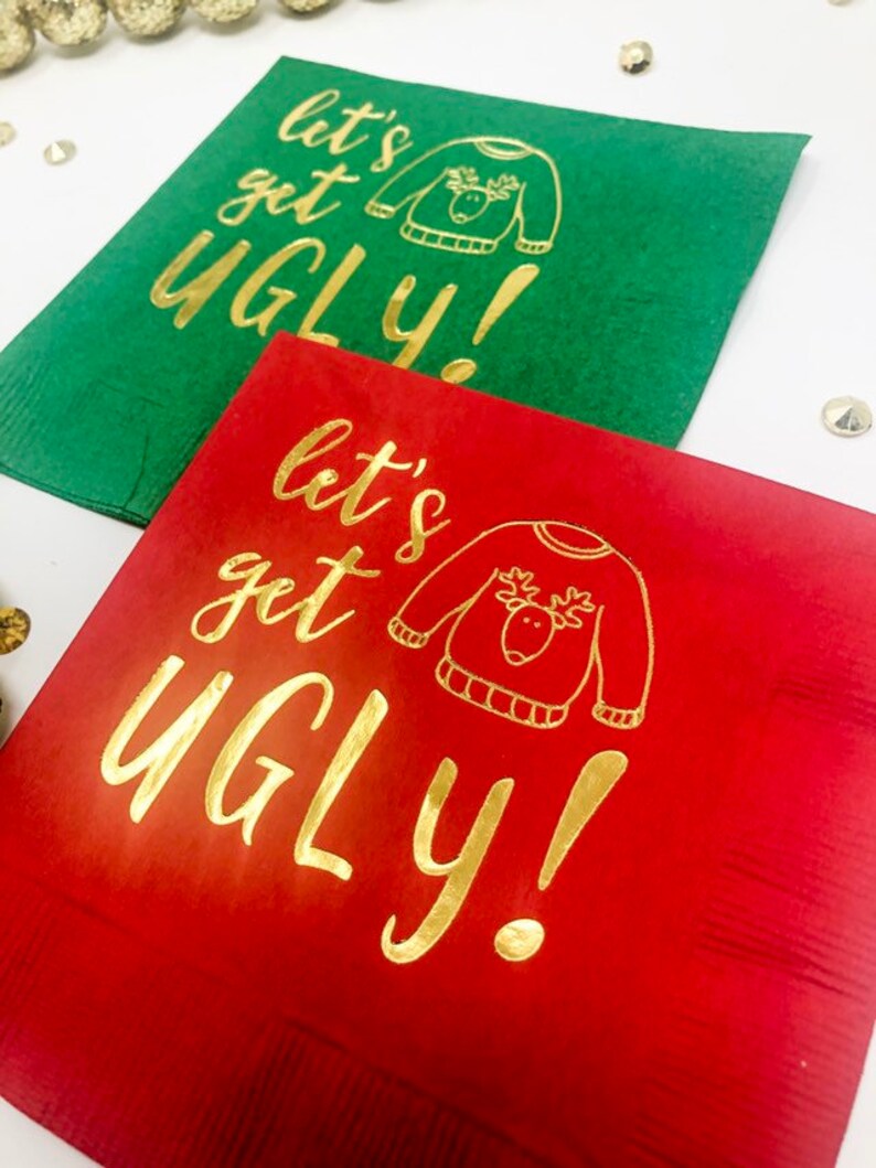 May include: Two square paper napkins, one green and one red, each featuring gold foil text that reads "let's get UGLY!" alongside a simple line drawing of a sweater with a reindeer face on the front, arranged on a white surface.