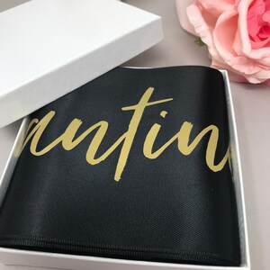 May include: A black satin pocket square with gold script text inside a white gift box. The pocket square is folded neatly, and the gold text is visible. The box is open, revealing the contents. Soft focus background with pink and white flowers.