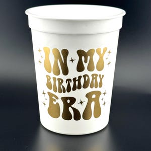 May include: White plastic cup with gold text that reads "IN MY BIRTHDAY ERA" in a retro font. The cup has a slightly flared rim and is decorated with gold starbursts. The cup is designed for a birthday celebration.