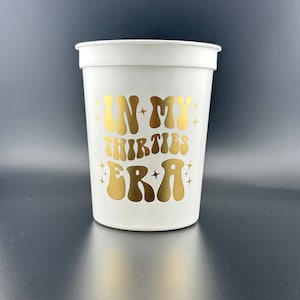 Pode incluir: Copo de pl&aacute;stico branco com um design dourado que diz "In My Thirties Era" com estrelas ao redor do texto.