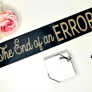 A black satin sash with "The End of an ERROR" in gold glitter script, next to a small white gift box and pink roses.