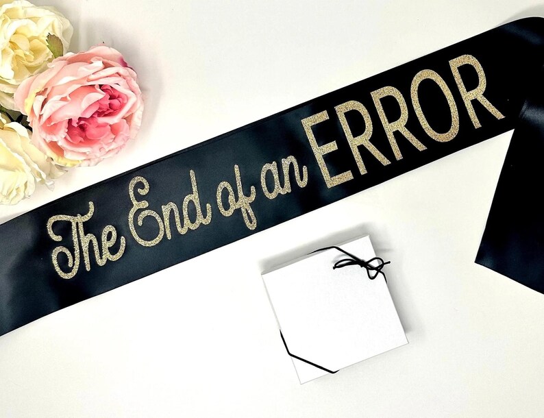 A black satin sash with "The End of an ERROR" in gold glitter script, next to a small white gift box and pink roses.