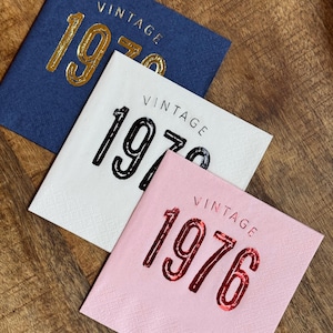 May include: Three square napkins with the word "VINTAGE" above the year. One is navy blue with gold lettering for "1970", another is white with black lettering for "1976", and the last is pink with red lettering for "1976".