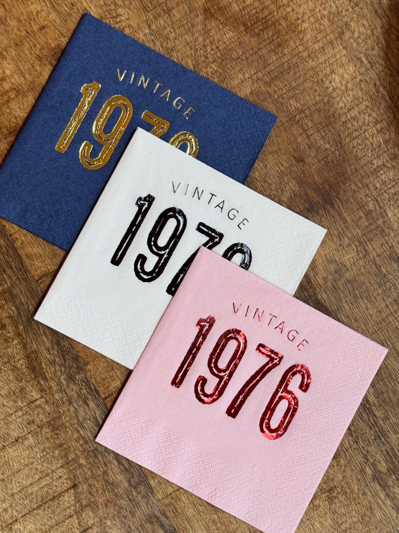 May include: Three square napkins with the word "VINTAGE" above the year. One is navy blue with gold lettering for "1970", another is white with black lettering for "1976", and the last is pink with red lettering for "1976".