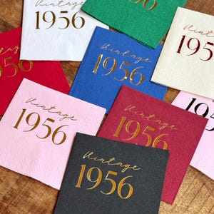 May include: A collection of square paper napkins in various colors, including white, red, pink, blue, green, and black. Each napkin is printed with the words "Vintage 1956" in gold script, making them suitable for a birthday celebration.