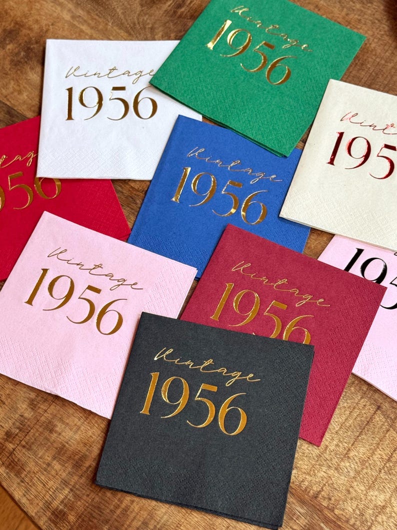 May include: A collection of square paper napkins in various colors, including white, red, pink, blue, green, and black. Each napkin is printed with the words "Vintage 1956" in gold script, making them suitable for a birthday celebration.