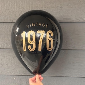 Puede incluir: Globo negro con texto dorado "VINTAGE 1976". El globo se sostiene frente a un fondo de madera gris. La superficie del globo es reflectante.