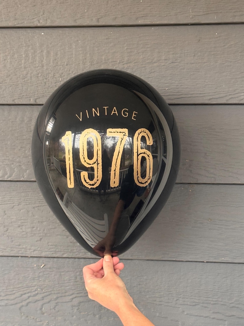 Puede incluir: Globo negro con texto dorado "VINTAGE 1976". El globo se sostiene frente a un fondo de madera gris. La superficie del globo es reflectante.