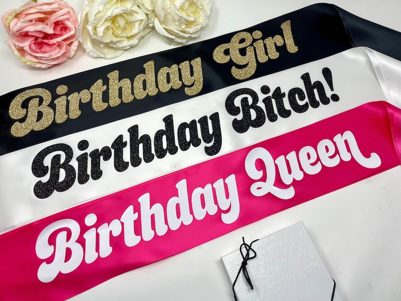 Three satin sashes with the text "Birthday Girl", "Birthday Bitch!" and "Birthday Queen" in different fonts and colors. The sashes are black, white, and pink.  The text is in a retro theme
