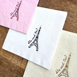 May include: Three paper napkins in pink, white, and cream, each with a printed Eiffel Tower design and the phrase "She said Oui!". The napkins are arranged on a wooden surface.