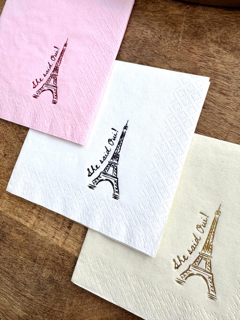 May include: Three paper napkins in pink, white, and cream, each with a printed Eiffel Tower design and the phrase "She said Oui!". The napkins are arranged on a wooden surface.