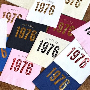 May include: A collection of square napkins in various colors, each printed with the word "VINTAGE" above the year "1976". The napkins feature gold, black, and red lettering on backgrounds of white, pink, navy, and burgundy.