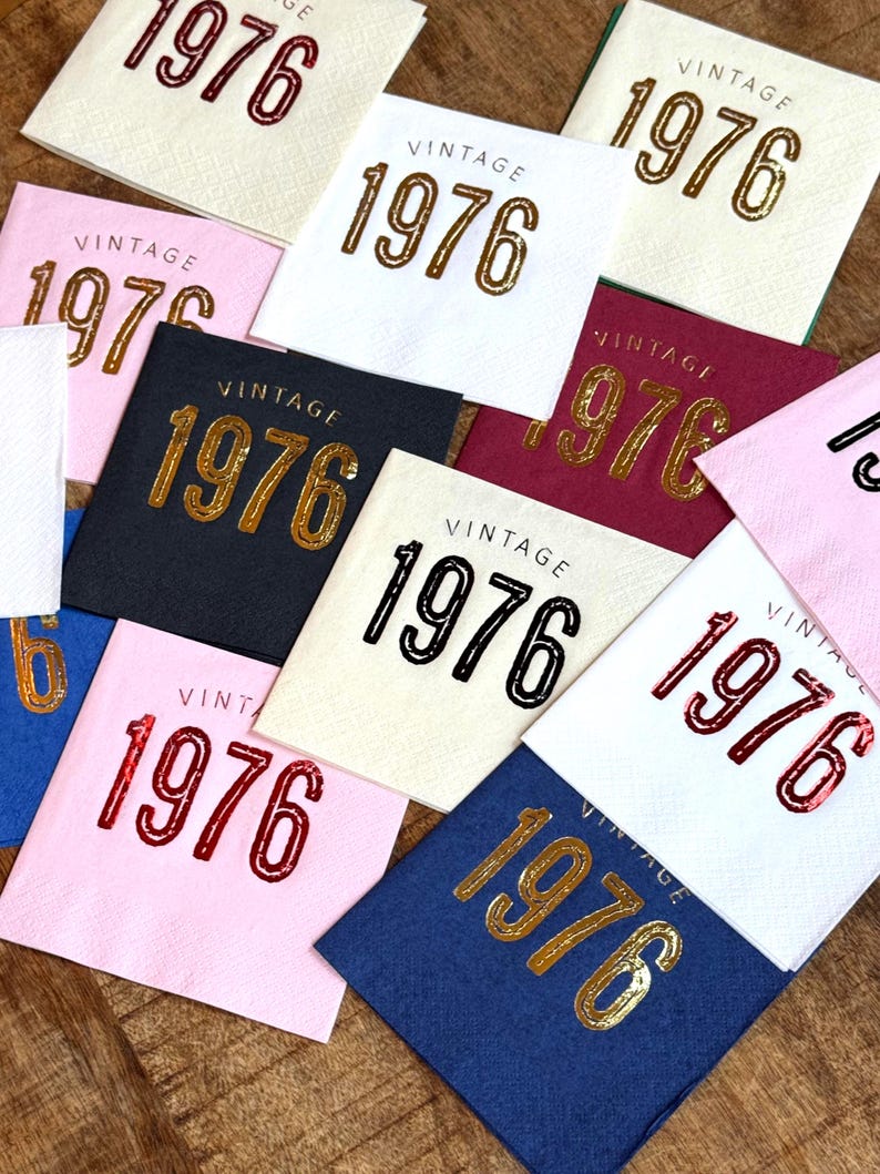 May include: A collection of square napkins in various colors, each printed with the word "VINTAGE" above the year "1976". The napkins feature gold, black, and red lettering on backgrounds of white, pink, navy, and burgundy.