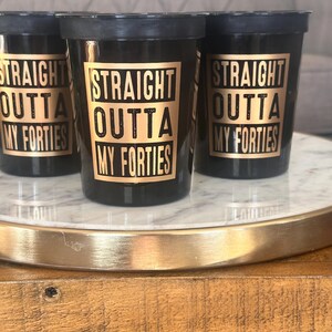 Three black cups printed with gold graphics reading Straight Outta My Forties set on a marble base.