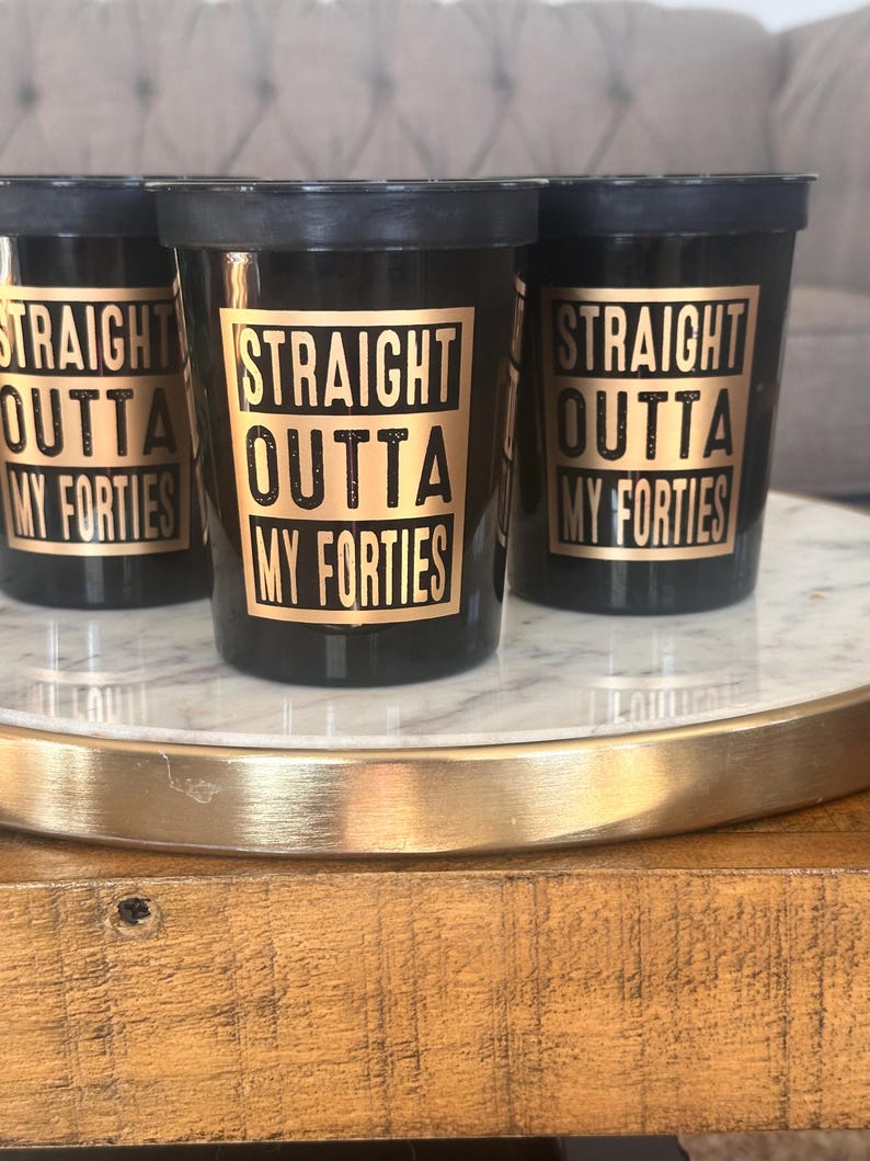 Three black cups printed with gold graphics reading Straight Outta My Forties set on a marble base.