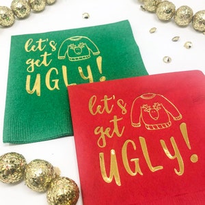 May include: A green paper napkin and a red paper napkin, each featuring gold foil text that reads "let's get UGLY!" alongside a line drawing of a sweater with a reindeer face. The napkins are surrounded by gold glittery decorative spheres.