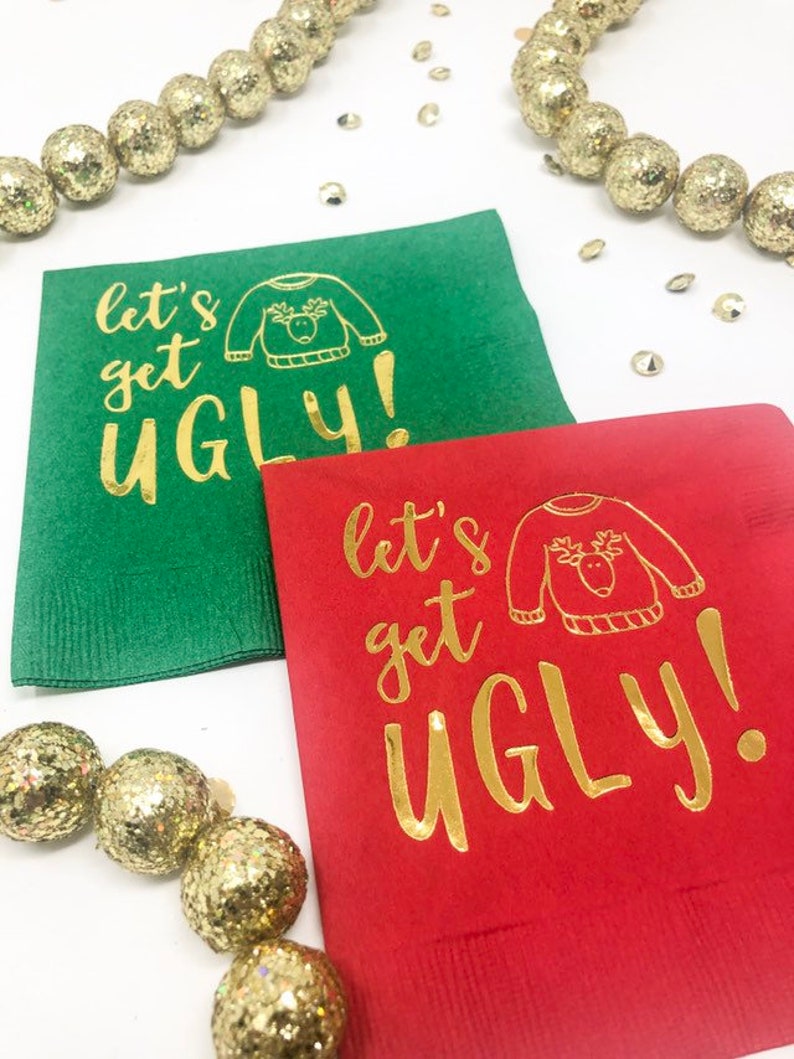 May include: A green paper napkin and a red paper napkin, each featuring gold foil text that reads "let's get UGLY!" alongside a line drawing of a sweater with a reindeer face. The napkins are surrounded by gold glittery decorative spheres.